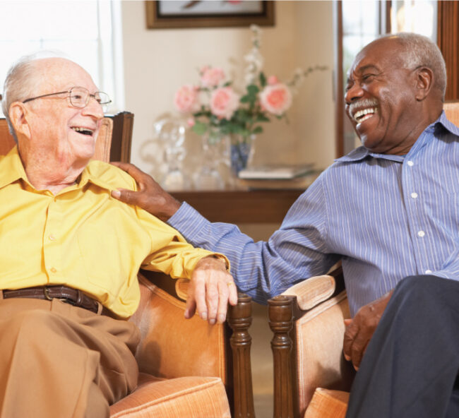 Assisted-Lifestyle Two senior men sitting in chairs and smiling while talking to each other