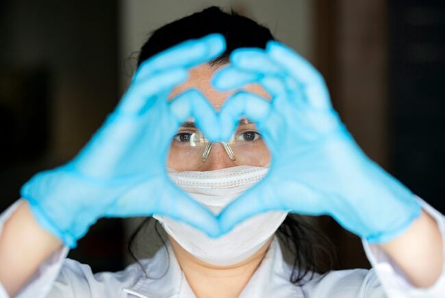 GettyImages-1219131671 Healthcare Worker with mask and blue gloves on her hand making the shape of a heart.