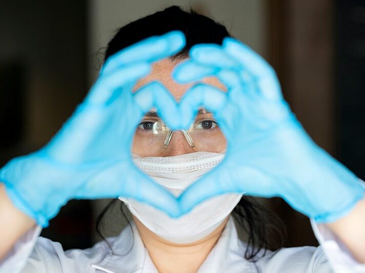 GettyImages-1219131671 Healthcare Worker with mask and blue gloves on her hand making the shape of a heart.